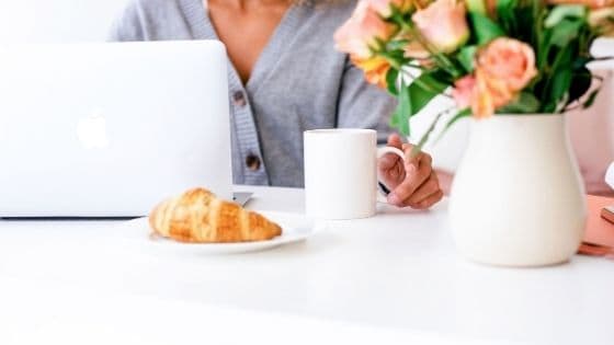 woman holding a white coffee mug next to her white case laptop and a white flower vase