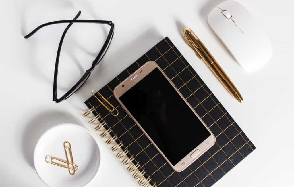 phone on a notebook next to a pen, white computer mouse and eyeglasses