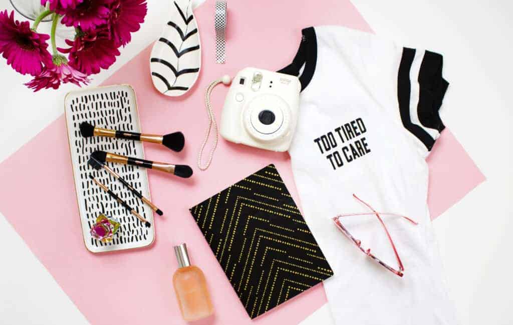 a photo of a white top next to a white camera, black notebook and some makeup brushes