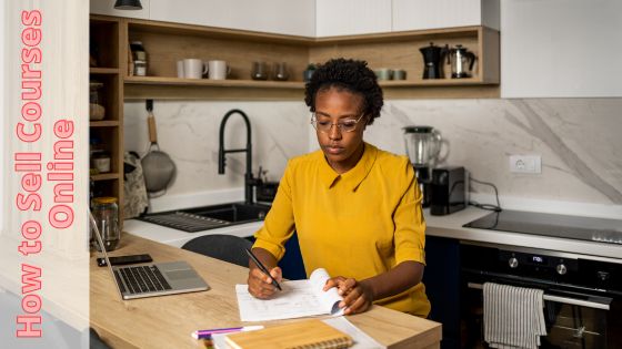 Photo of a woman sitting in a kitchen, writing in a book with an open laptop on the counter next to her